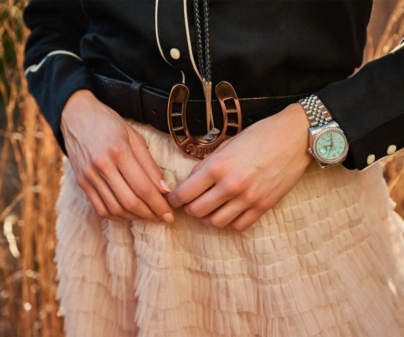 Close-up of a person's hands adjusting a belt with a horseshoe buckle and wearing a stylish watch, paired with a ruffled skirt.