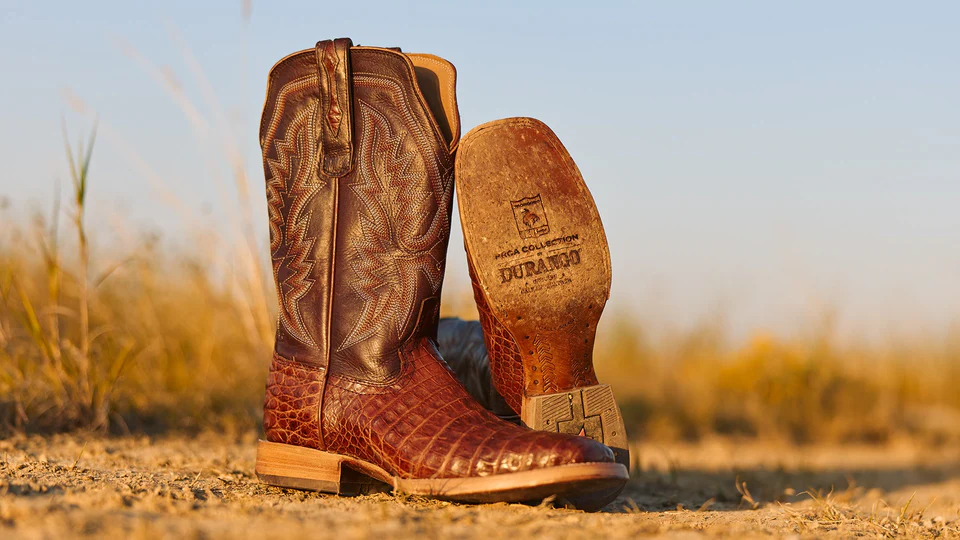 A pair of brown cowboy boots with intricate stitching, displayed on a dirt surface against a natural background.