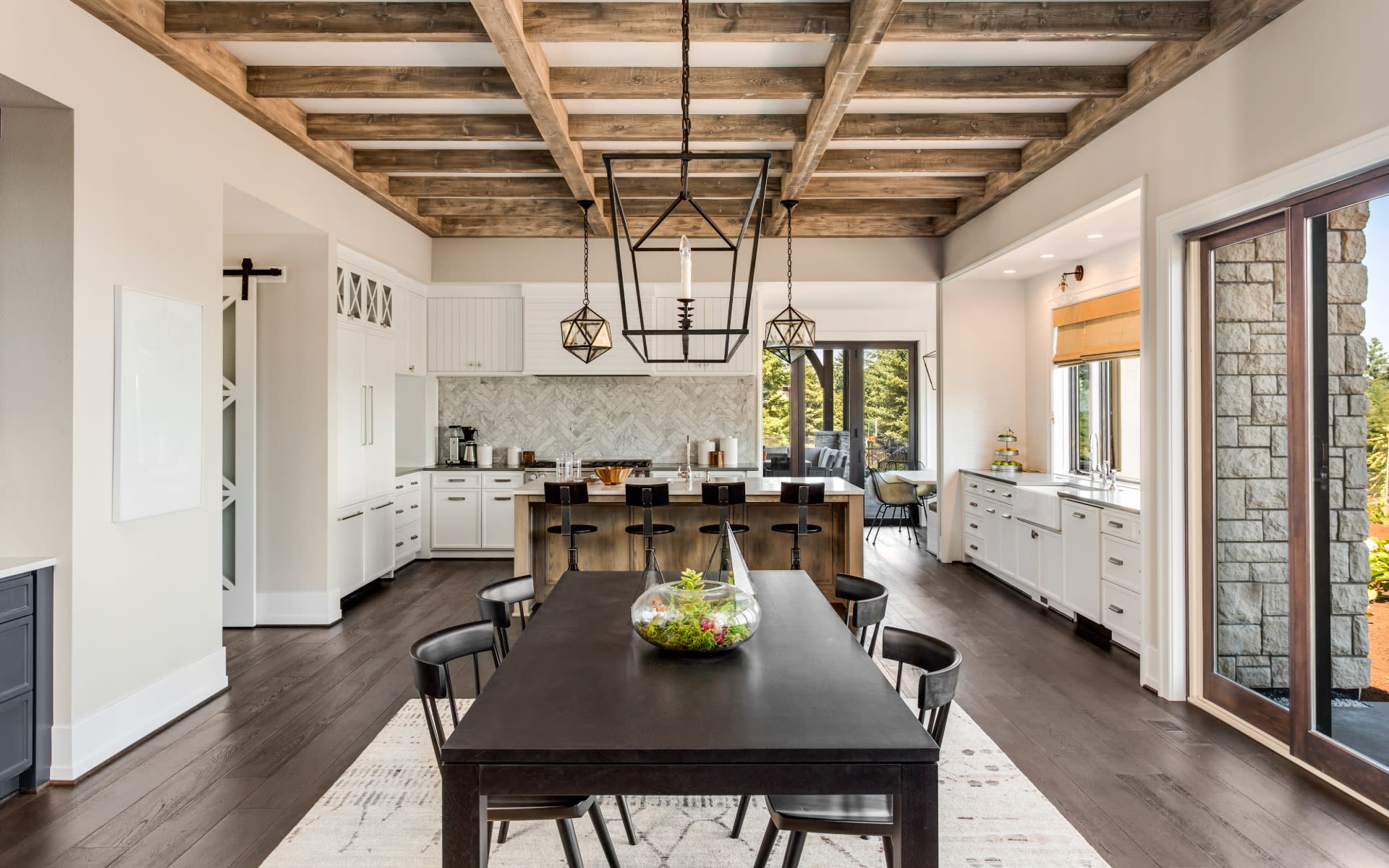 A modern kitchen featuring wooden beams, white cabinetry, and a central dining table with a glass planter. Three pendant lights hang above the dining area, with large windows providing natural light and a view of the outdoors.